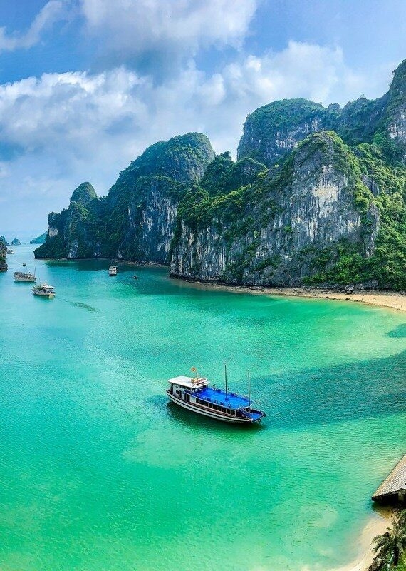 Aerial view of a boat in turquoise water surrounded by forested limestone mountains in Halong Bay.