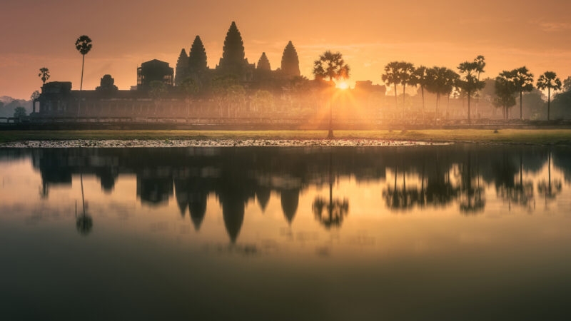 The sun rising behind the stone towers of Angkor Wat with a clear reflection in the water at the foreground.