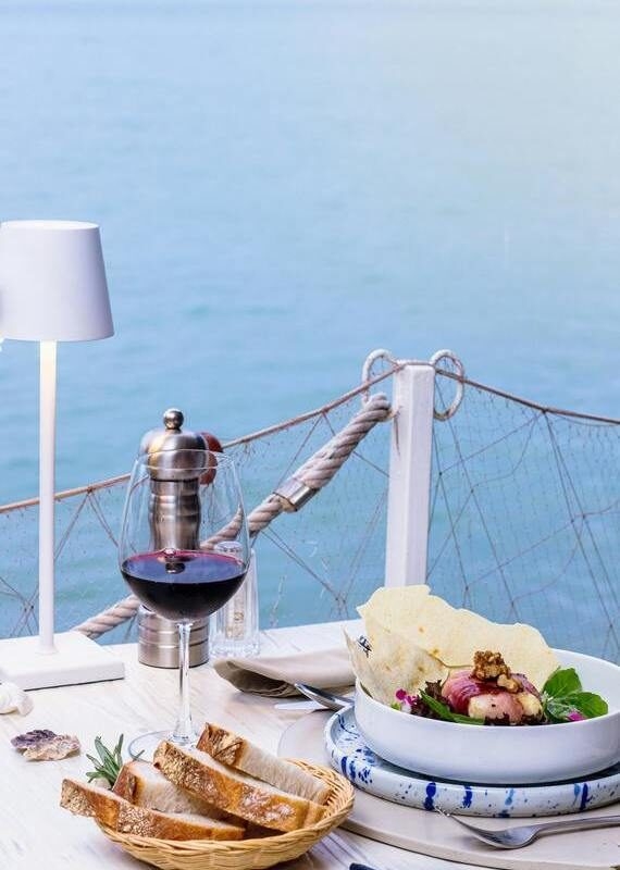 Gourmet meal with red wine and sliced bread on a table by the ocean at a seaside restaurant.
