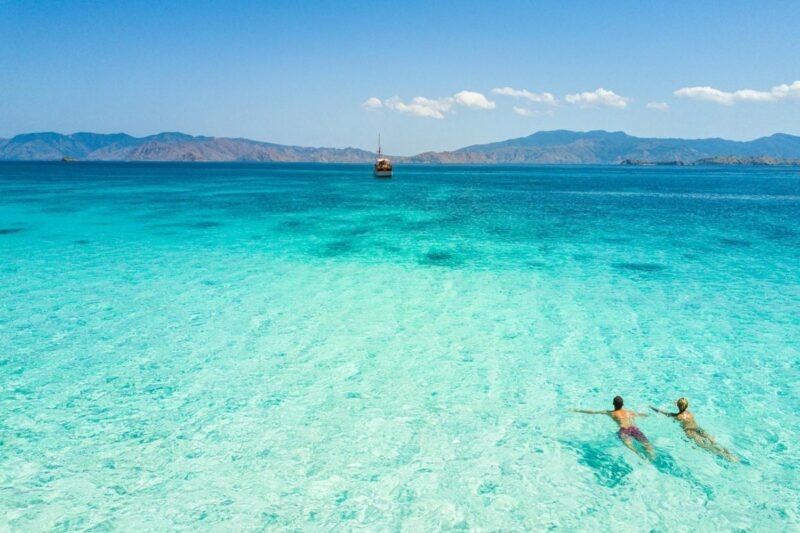 Two people swimming in clear tropical water toward a wooden boat near distant islands.