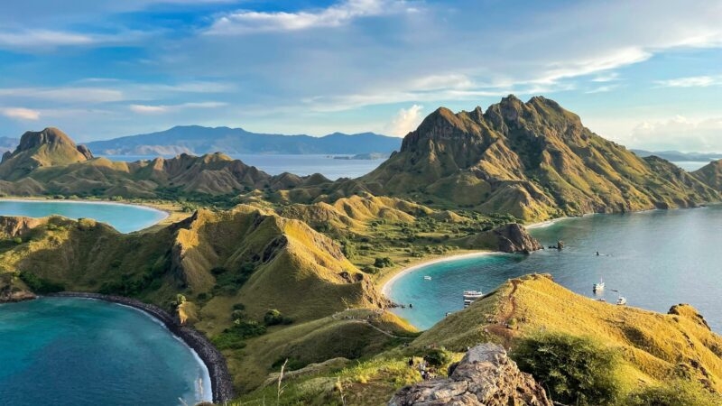 Landscape view of Padar Island with rolling green hills and multiple blue ocean bays under a cloudy sky.