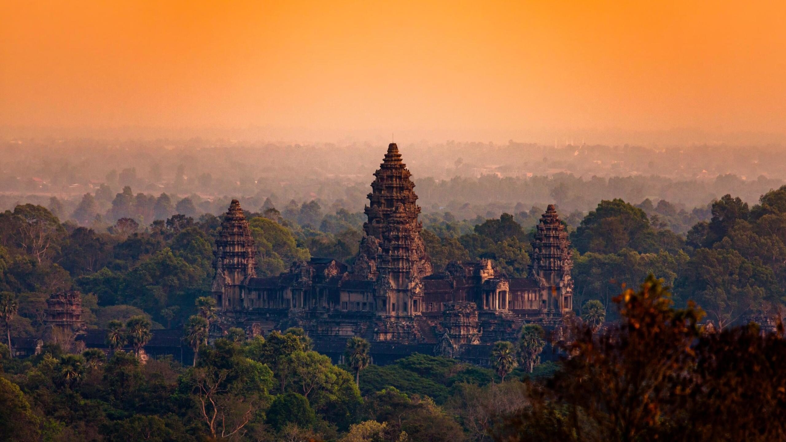 The silhouette of Angkor Wat's stone towers rising above a lush green forest under a glowing orange sunset sky.