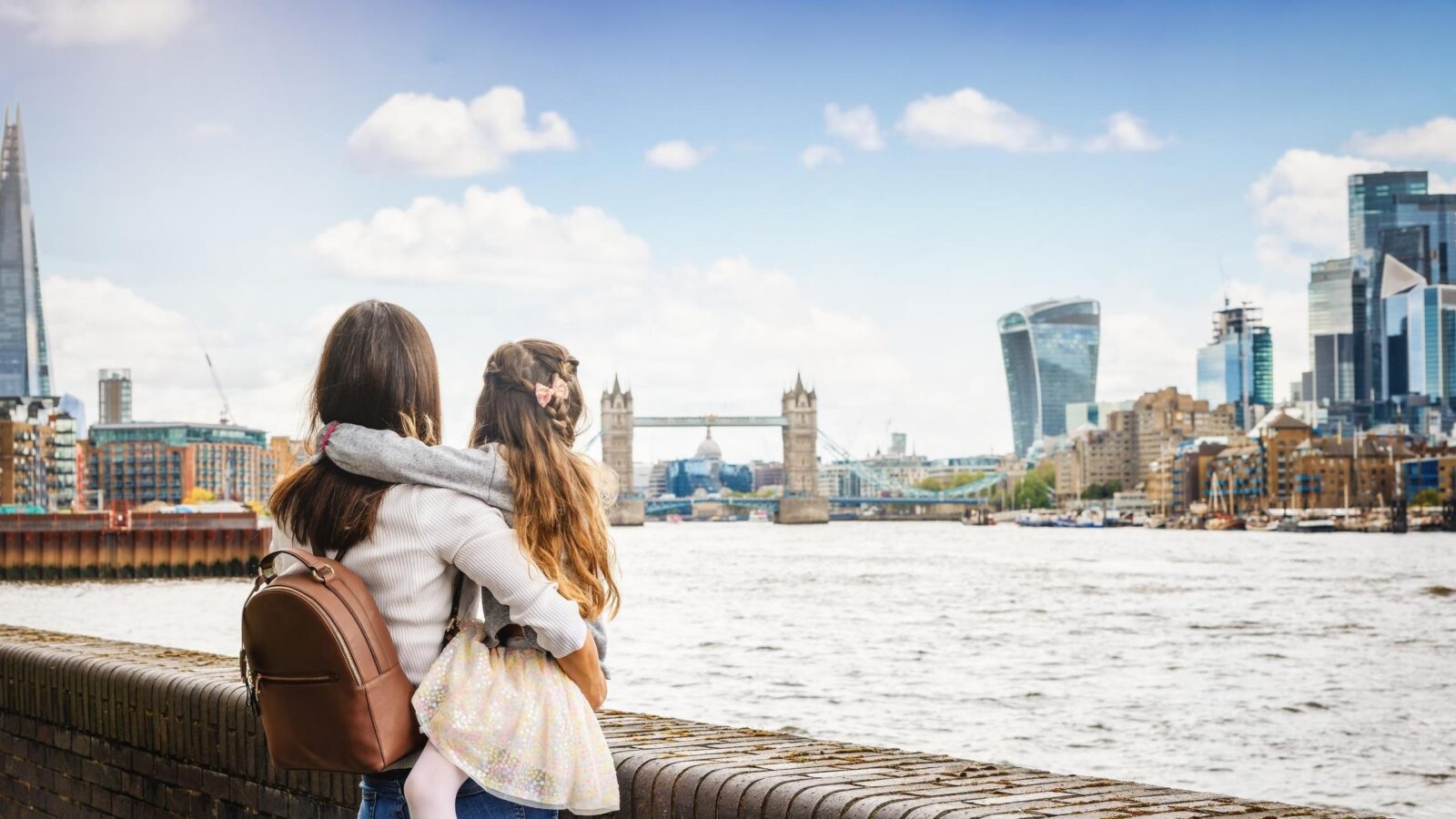 A woman carrying a young girl looks toward Tower Bridge and the city skyline from the banks of the River Thames.