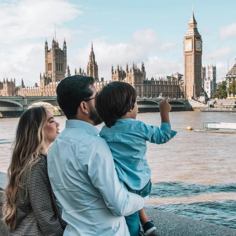 A man holds a child pointing toward Big Ben and the Houses of Parliament from across the river.