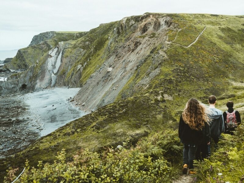 Three people hike along a green cliffside path overlooking a rugged coastline and rocky shore under an overcast sky.