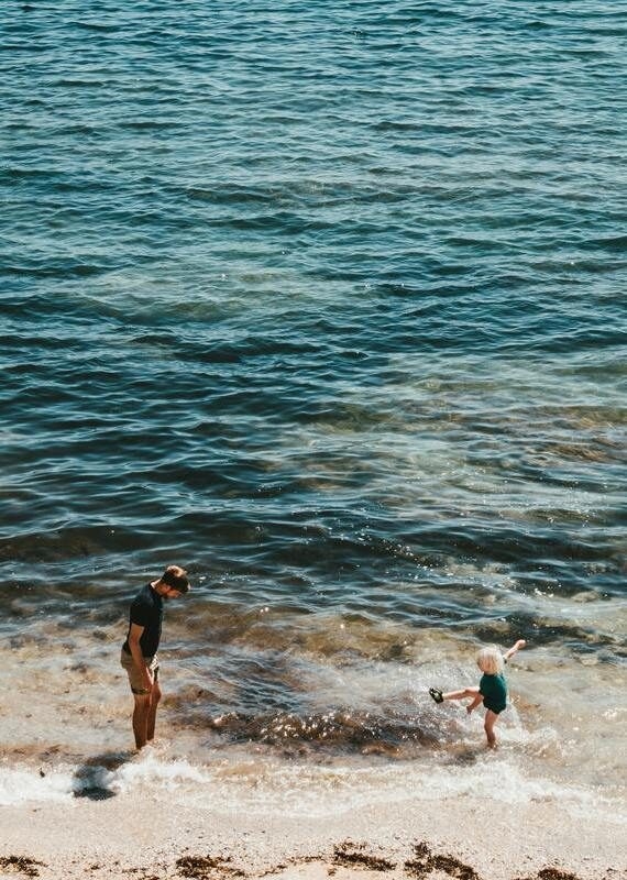 A man stands on a sandy shore watching a small child kick at the water's edge in the bright sunlight.