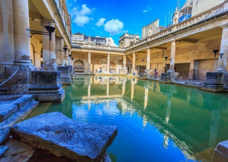 Close-up of steaming green water in a Roman bath surrounded by stone pillars and lit torches.