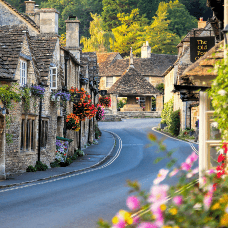 A quaint English village street with stone cottages, floral displays, and "The Old Post Office" sign.
