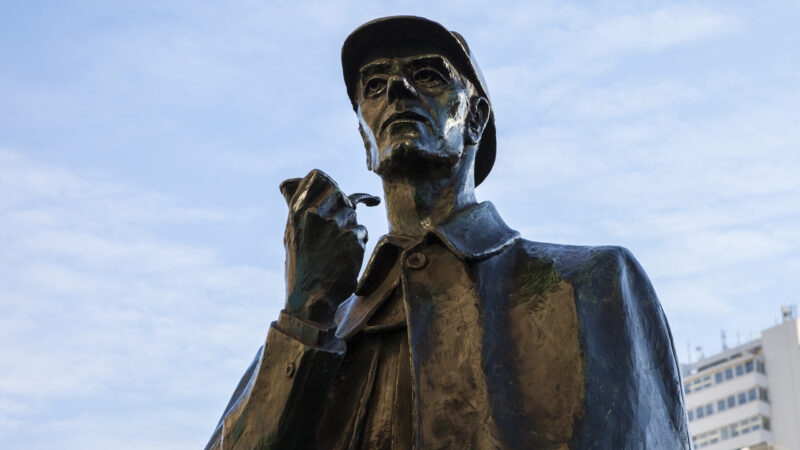 Bronze statue of Sherlock Holmes in a deerstalker hat holding a pipe against a bright sky.