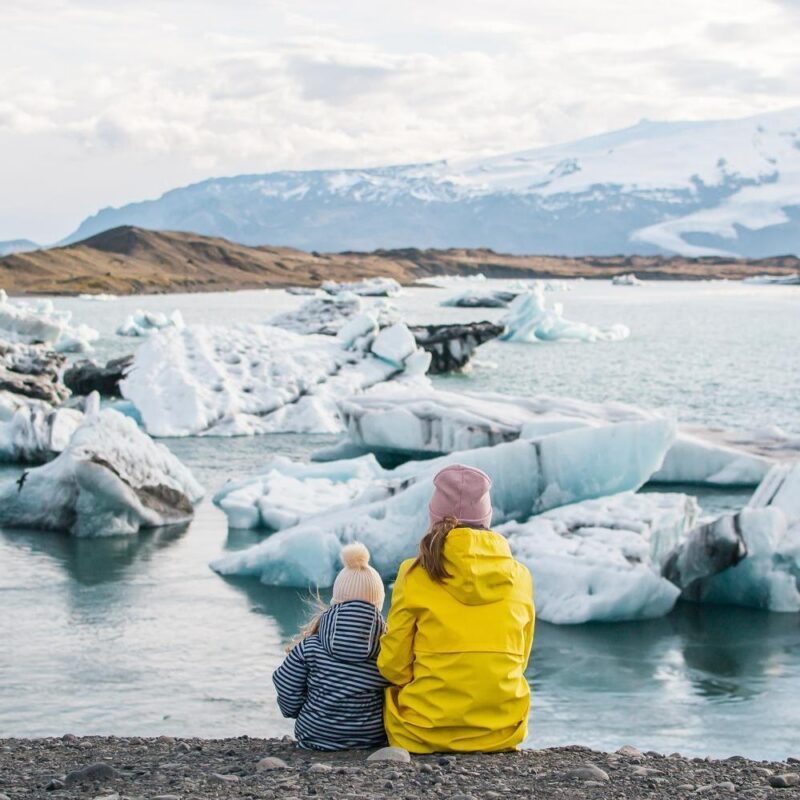Mother and child in warm coats sitting by a lagoon filled with icebergs on an Iceland family vacation.