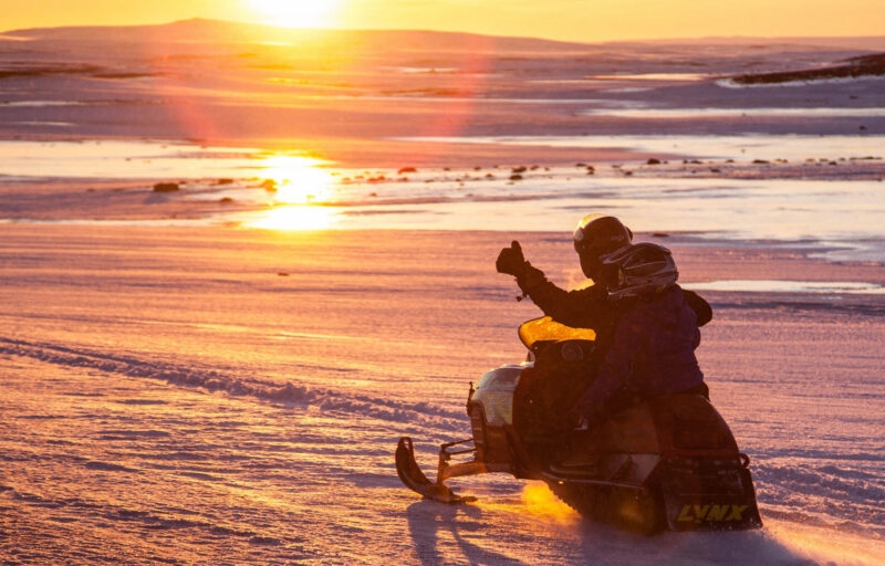 Two people riding a snowmobile on a snowy field at sunset during an Iceland family vacation.