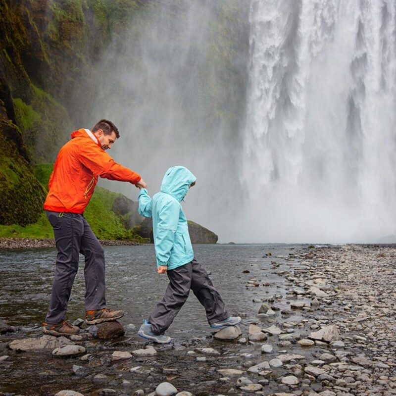 A father and child hiking across river stones at the base of a powerful waterfall on an Iceland family vacation.
