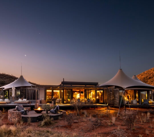 Exterior view of a tented camp at dusk, with three pointed tents surrounding a glass and open-air lobby with a large deck and in the foreground a table, chairs and firepit, all set in bushland with red soil and short grass.