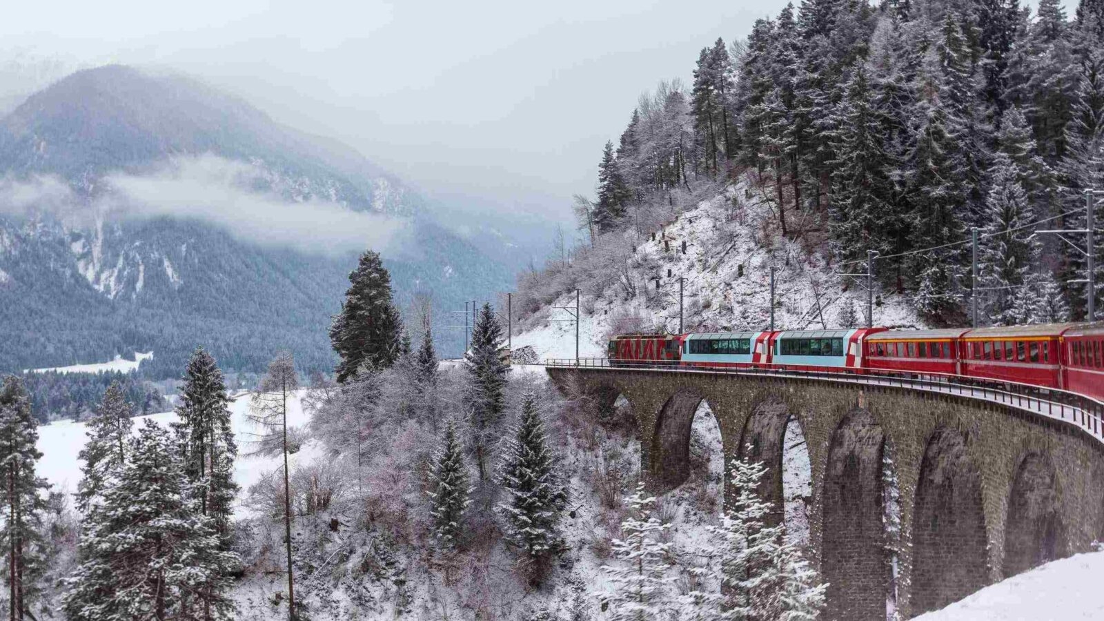 A red passenger train crosses a tall stone viaduct bridge in a snowy mountain forest in Switzerland.