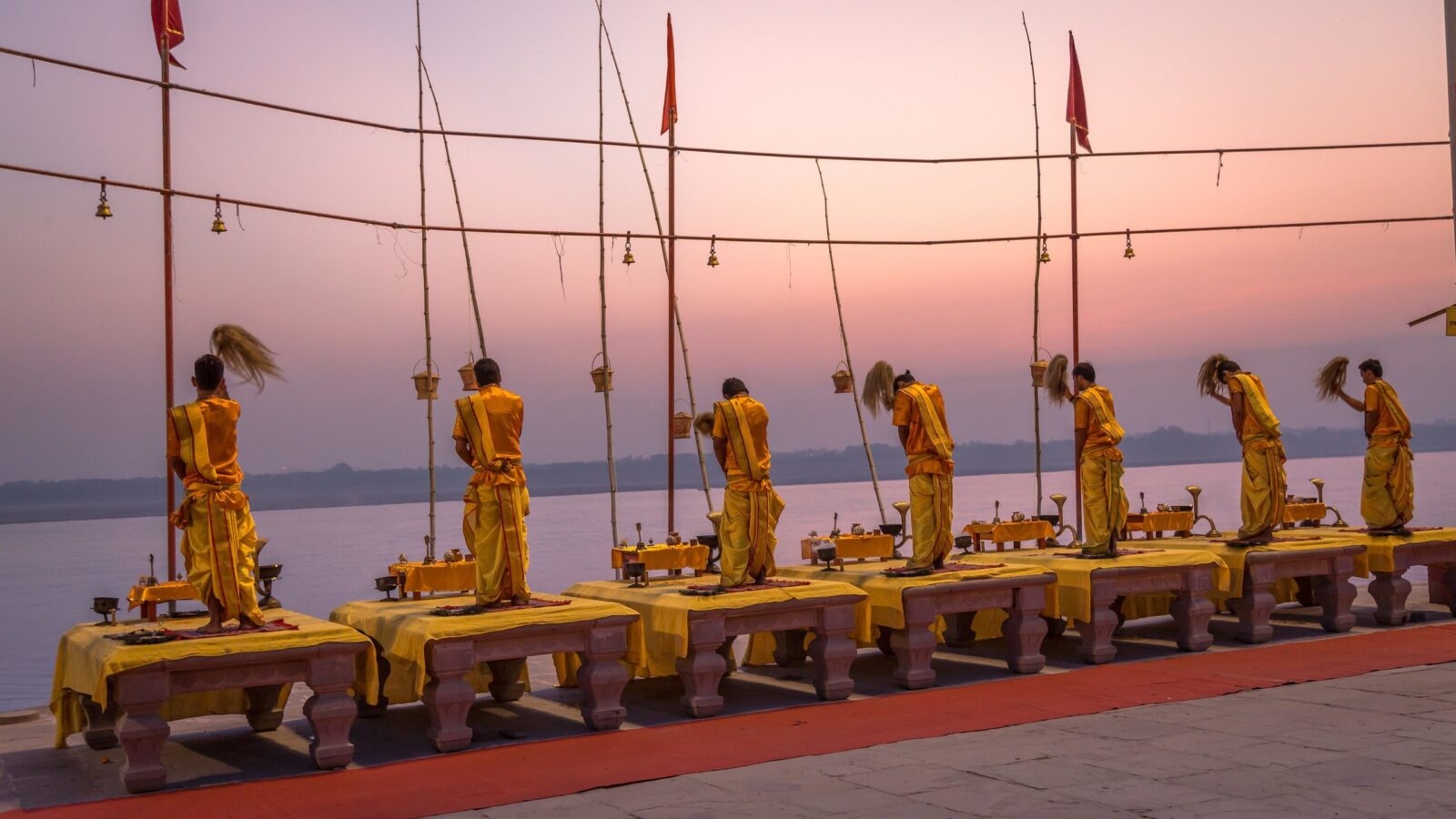 Priests in yellow robes perform a ritual with whisks on platforms overlooking a wide river during a pink sunset.