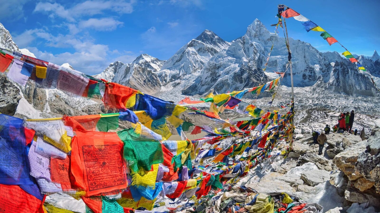 Colorful prayer flags stretch across a rocky mountain pass with massive snow-covered Himalayan peaks in the background.