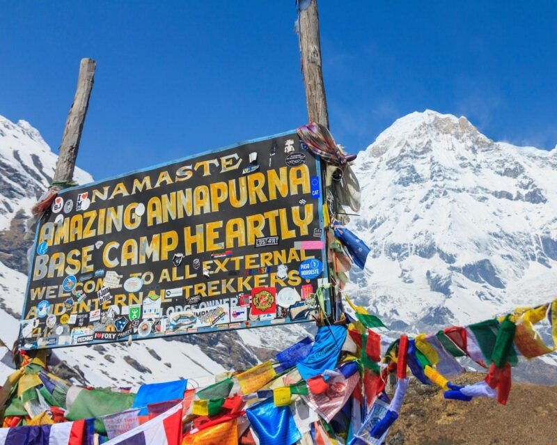 A sign reads Namaste Amazing Annapurna Base Camp Heartly Welcome, covered in stickers with snowy mountains behind it.