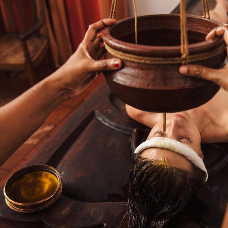 Warm herbal oil pours from a clay pot onto a person's forehead during a traditional Shirodhara Ayurvedic treatment.