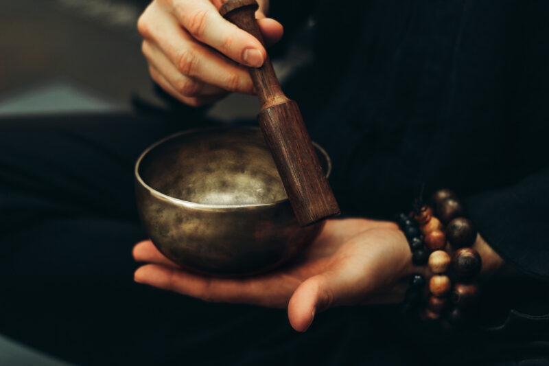 Close-up of a person holding a bronze singing bowl and wooden mallet, wearing beaded bracelets during a sound bath.