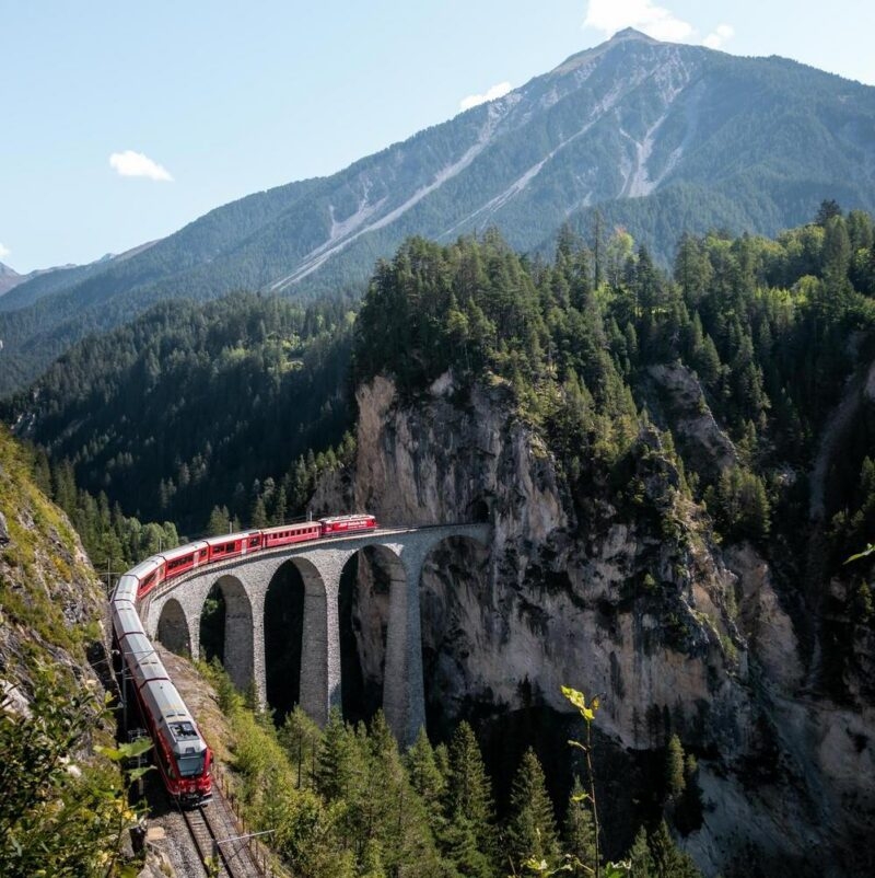 A red train crossing a high arched stone viaduct over a deep forest ravine toward a mountain tunnel.