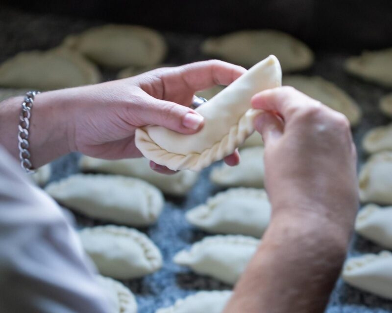 Close-up of hands folding the decorative edge of a raw empanada over a table of prepared pastries.