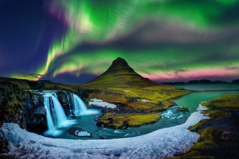 A long exposure shot of a waterfall and the Kirkjufell mountain under a colorful green and pink aurora sky.
