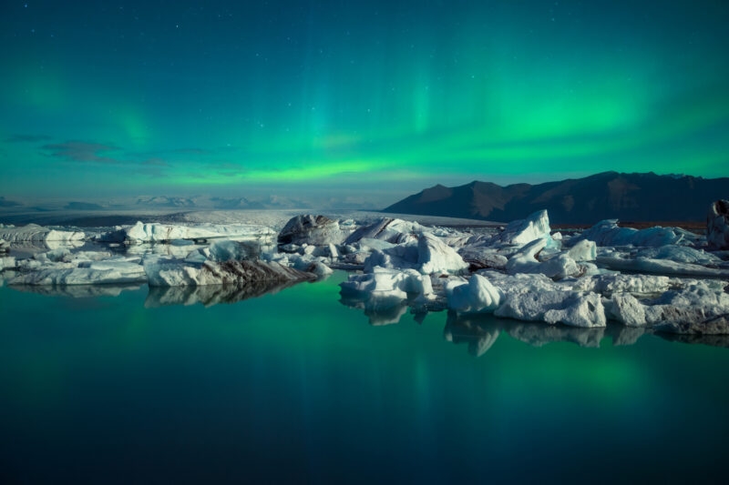 Green northern lights illuminate the night sky above a lagoon with icebergs and distant mountains in Iceland.