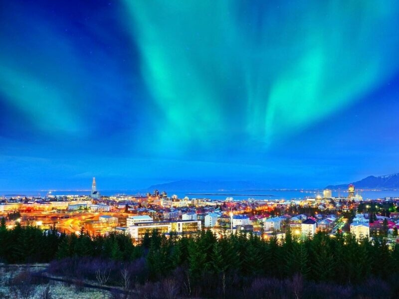 Bright blue and green northern lights stretch across the sky over the illuminated cityscape of Reykjavik, Iceland.