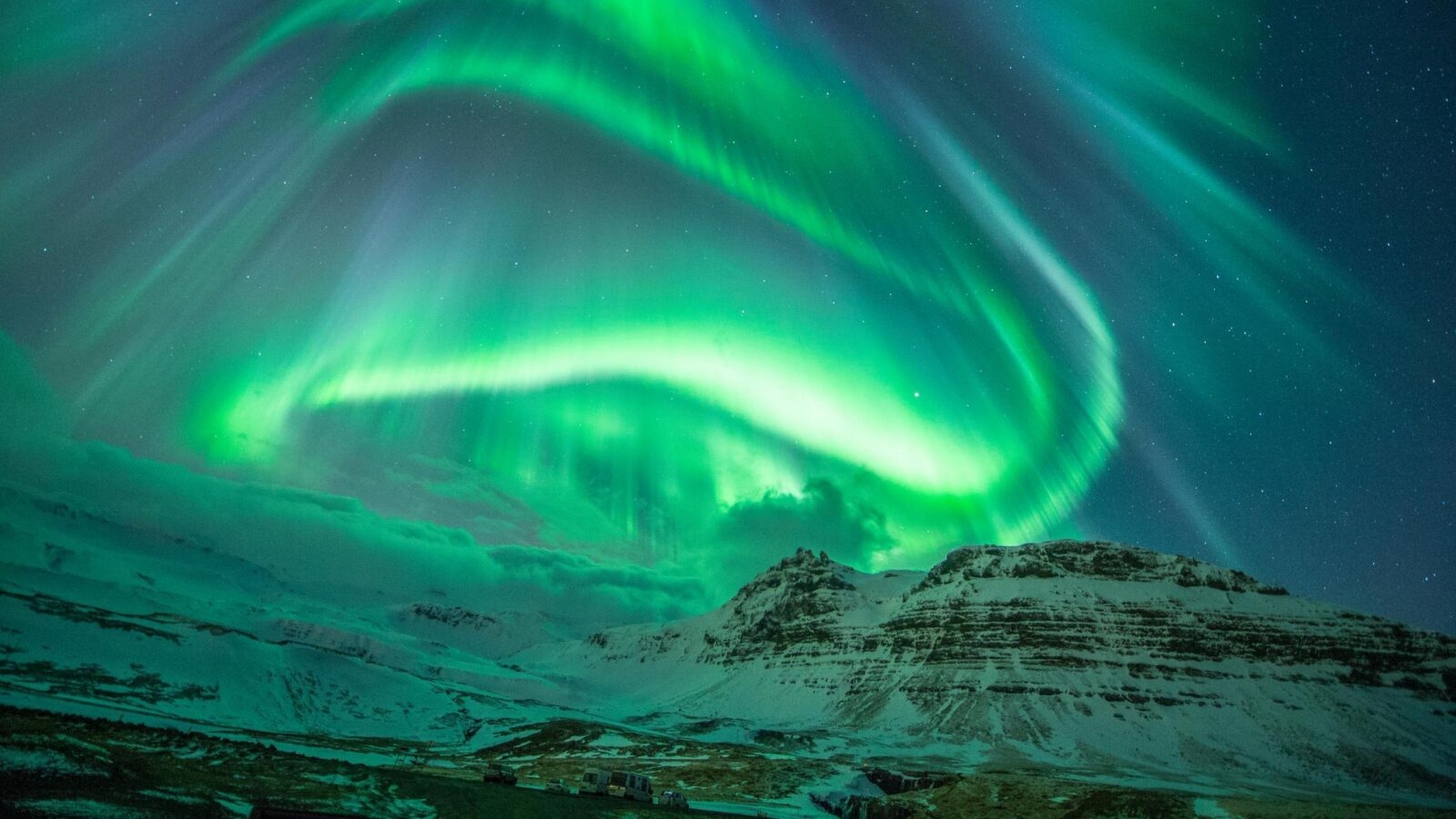 Swirling green aurora borealis lights dance in the night sky over a snowy mountain landscape in Iceland.
