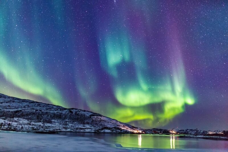 Green and purple aurora borealis over a snowy mountain and frozen water reflecting lights from a distant shore.