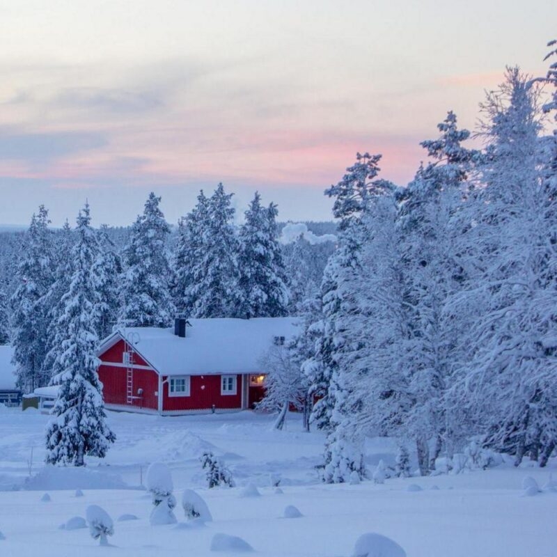 A red cabin surrounded by heavy snow and frost-covered trees under a pink and purple twilight sky in Finnish Lapland.