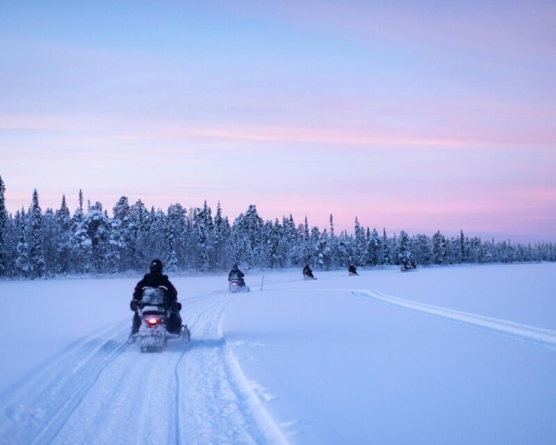People riding snowmobiles in a row across a deep snow field toward a line of frosted trees during a pink sunset.