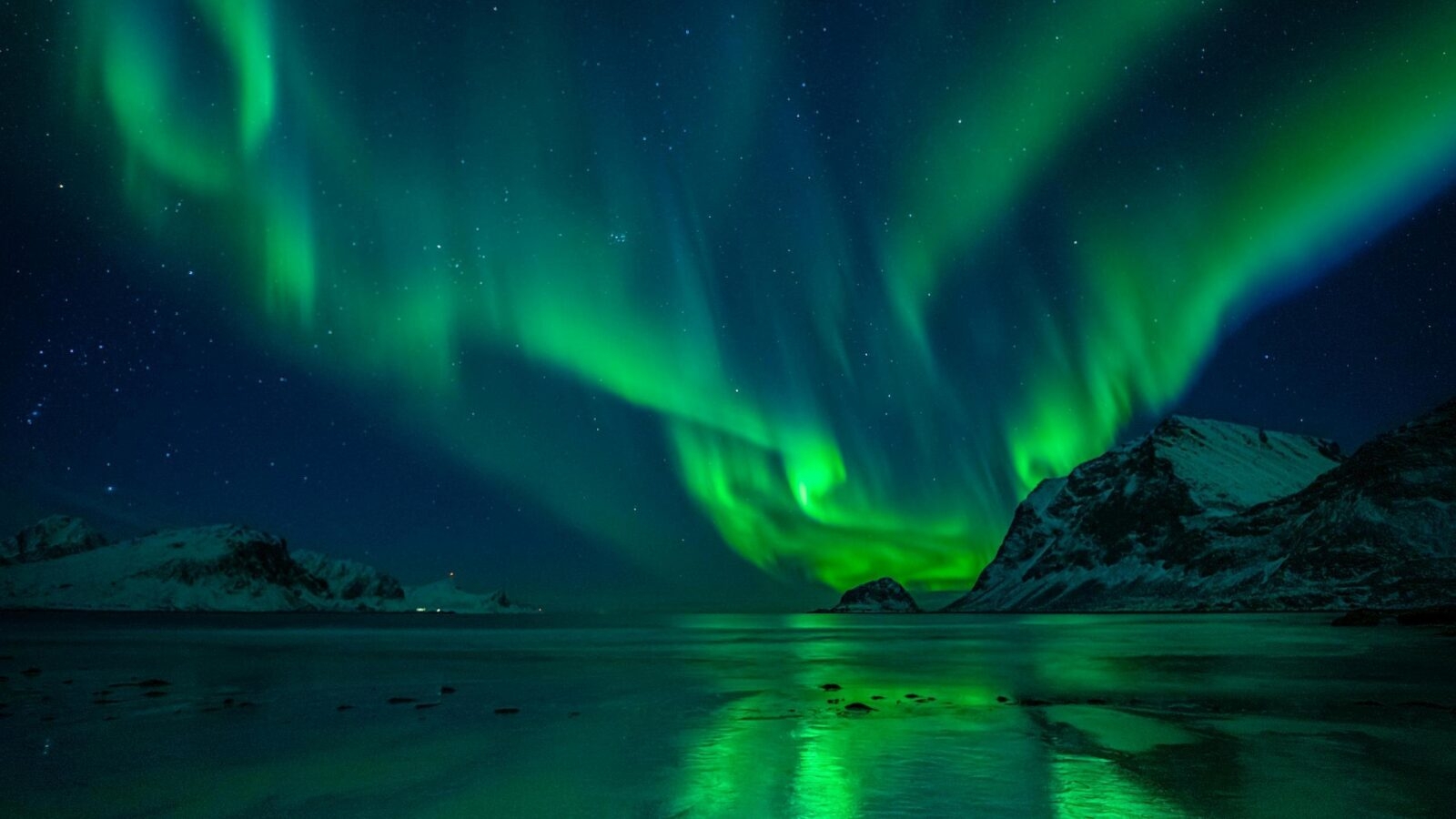 Green northern lights illuminate a starry night sky above snowy mountains and a reflective body of water.