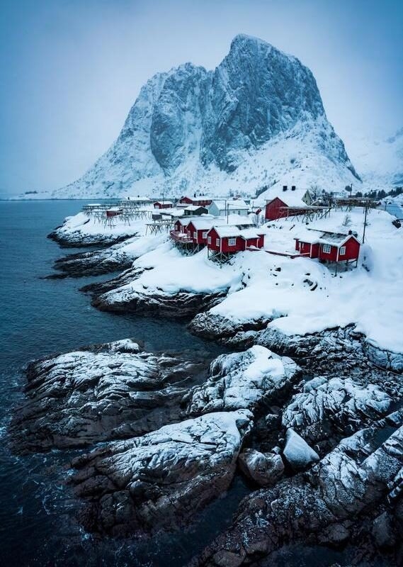 Red wooden houses on a snowy coastline in front of a massive, steep mountain peak under a cloudy sky.