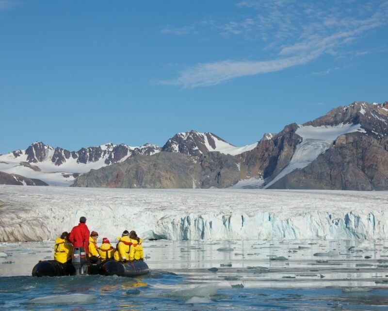 People in an inflatable boat navigate icy waters in front of a large glacier and jagged mountains.
