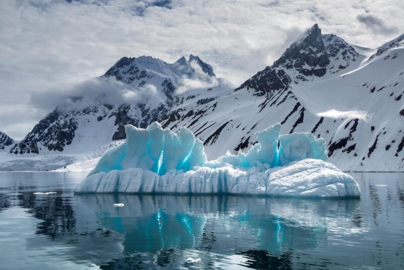 A large, translucent blue iceberg floating in calm water with snow-covered mountains in the background.