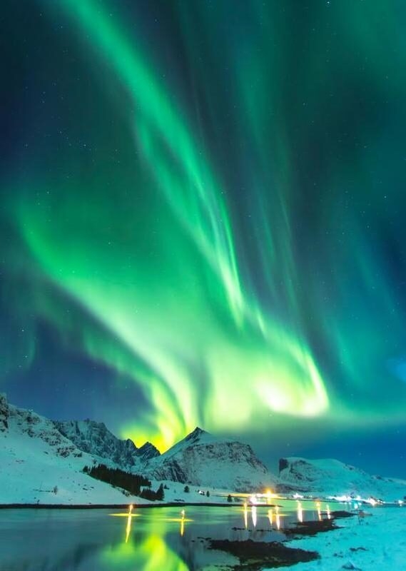 Vibrant green northern lights Norway stretching across a dark sky above snow-capped mountains and a calm harbor.