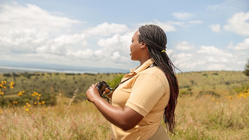 A woman holding binoculars looks out across an open, rolling green landscape under a blue sky with white clouds.