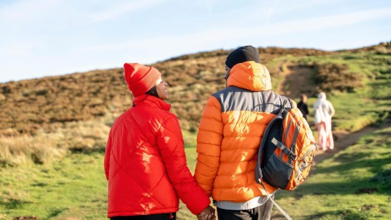 Rear view of a couple in red and orange jackets holding hands while hiking up a grassy hillside.