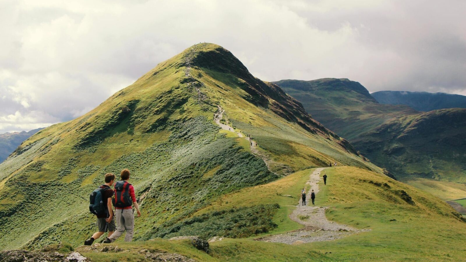 Two hikers with backpacks walking along a mountain ridge path leading toward a sharp green peak.