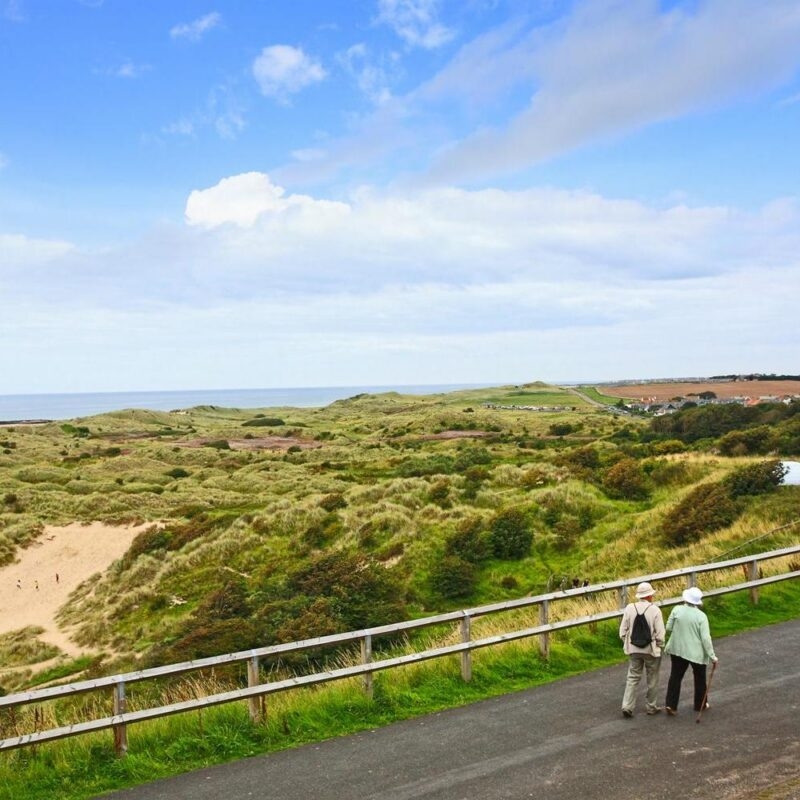 Older couple walking on a paved coastal path next to rolling green sand dunes and the distant sea.