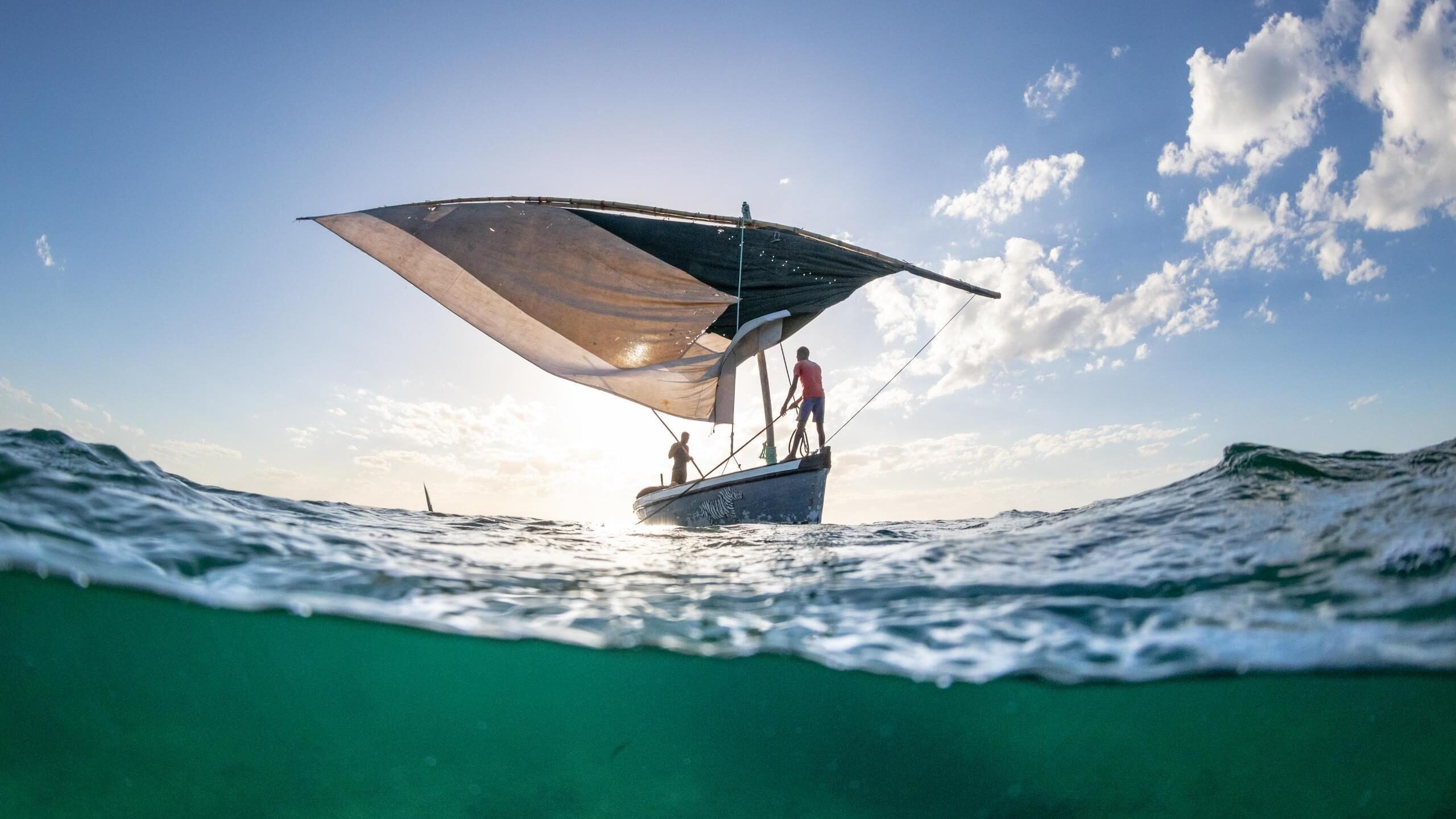 A split underwater and surface view of a traditional dhow boat sailing on clear turquoise ocean waters.