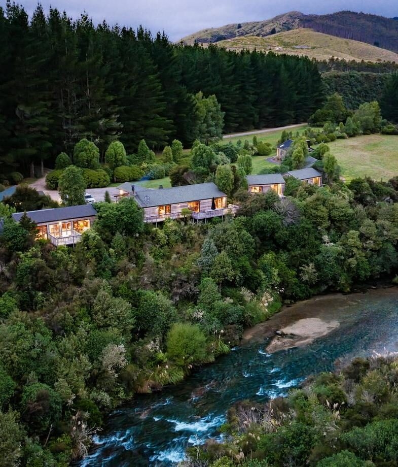 An aerial view of Poronui Lodge’s riverside cabins, followed by the peaceful pool terrace at Hapuku Lodge. Two secluded New Zealand stays surrounded by forest, water and mountain scenery.