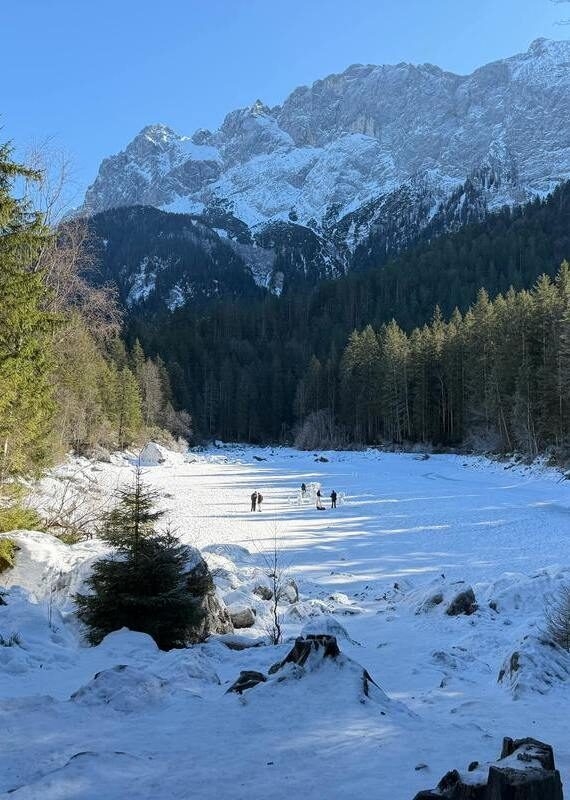 A snowy alpine valley in Bavaria with small figures walking near a pine forest under majestic winter mountains.