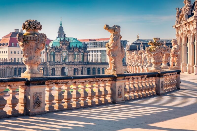 Classic stone statues and ornate urns line a grand terrace overlooking historic baroque buildings in Dresden, Germany.