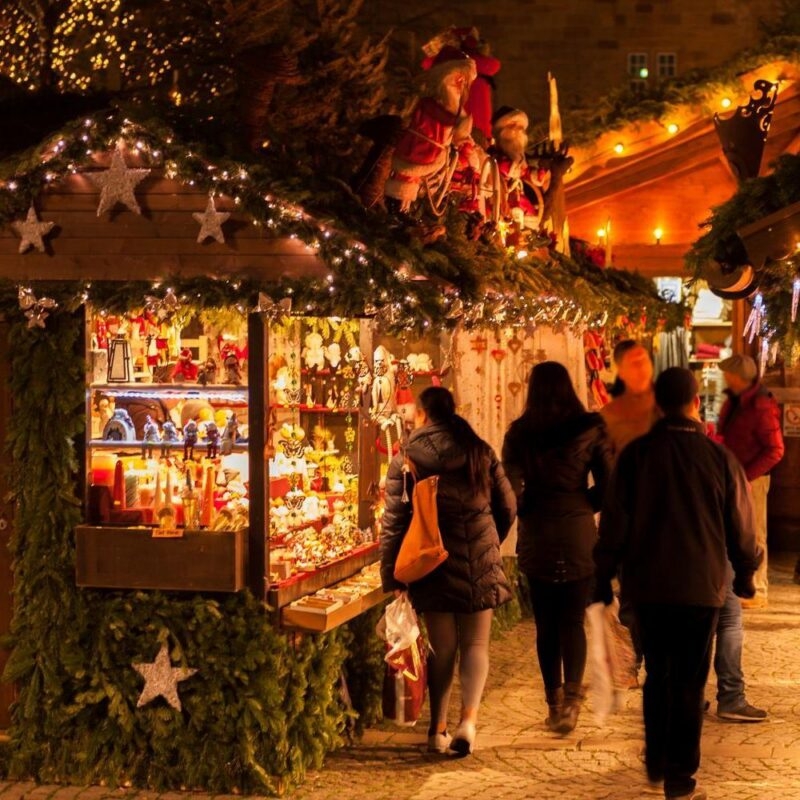 People browsing a brightly illuminated Christmas market stall filled with festive ornaments and decorations at night.
