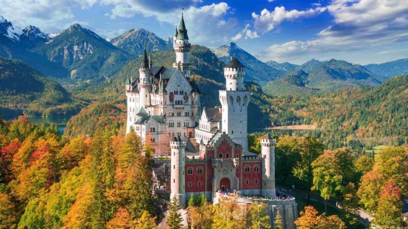Neuschwanstein Castle surrounded by colorful autumn trees with mountains in the background.