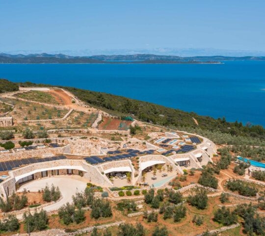 Panoramic aerial view of Villa Nai 3.3 built into a green coastal hillside under a clear blue sky.