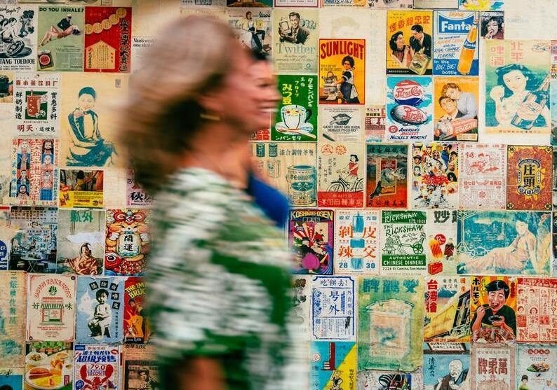 A mother and daughter on a trip exploring Bangkok's Chinatown walk past a wall adorned with vintage posters.