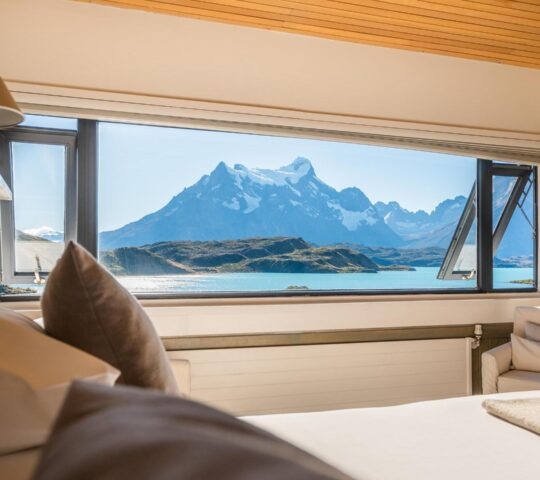 Large hotel window displaying a view of mountains and a bright blue lake from a bed in Explora Patagonia.